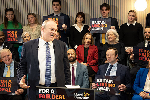 Ed Davey making a speech, a crowd of people are in the background holding signs reading "Britain Leading Again" and "For a Fair Deal"